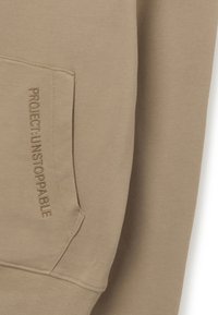 Sudadera beige con un bolsillo tipo canguro, puños acanalados y texto bordado "PROYECTO: INDETENIBLE" en el bolsillo frontal.