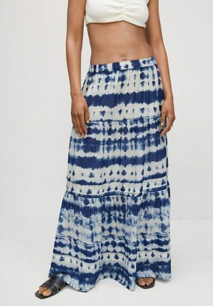Femme portant une jupe maxi tie-dye bleue et blanche, associée à des sandales noires et un top court blanc, debout devant un fond uni.