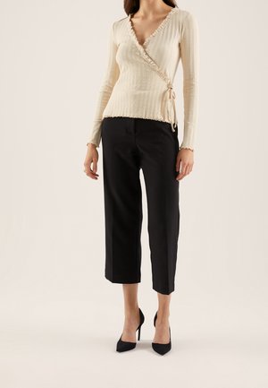 Frau trägt einen cremefarbenen, gerippten Wickelpullover mit Rüschenbesatz, schwarze knöchellange Hose und schwarze spitze High Heels, steht vor neutralem Hintergrund.