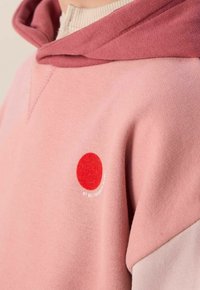 Hoodie rose avec un design stratifié et une capuche rose plus foncée. Un patch circulaire rouge avec un texte blanc apparaît à l'avant. Texture douce.