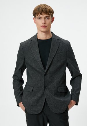 Schwarz- und graumeliertes Blazer mit Revers, zwei Knöpfen und Klappentaschen, getragen über einem schwarzen Rundhalsausschnitt-Shirt.