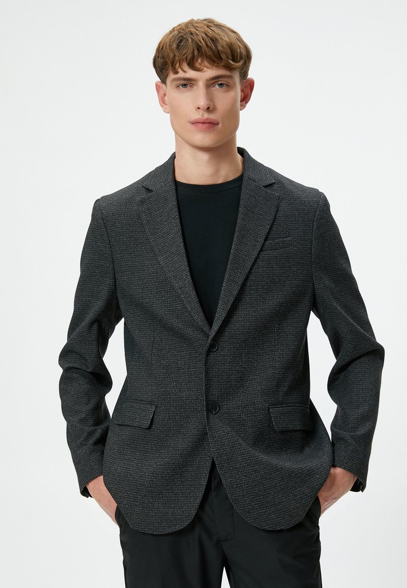 Schwarz- und graumeliertes Blazer mit Revers, zwei Knöpfen und Klappentaschen, getragen über einem schwarzen Rundhalsausschnitt-Shirt.