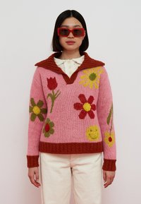 Knit sweater in pink with a brown ribbed collar, featuring floral patterns in green, red, and yellow. Textured fabric with a relaxed fit.