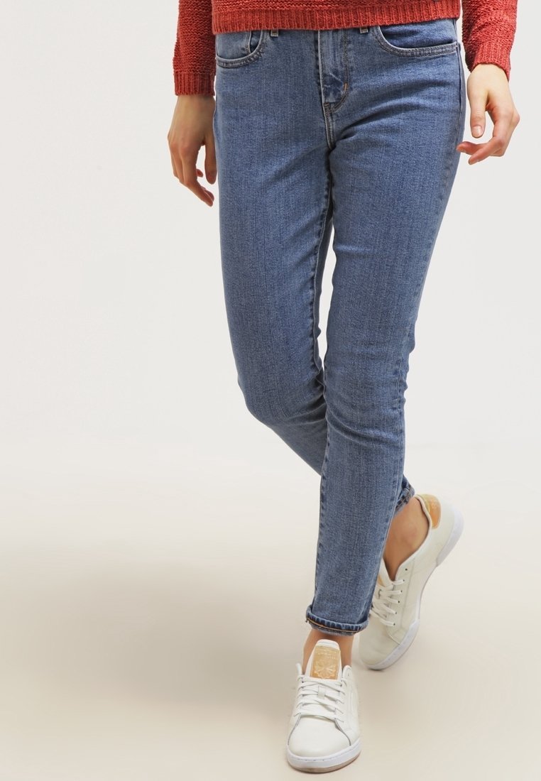 Schlanke blaue Jeans mit mittelhohem Bund, fünf Taschen und leichter Waschung. Kombiniert mit weißen Sneakers mit einem beigen Akzent.