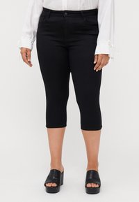 Black capri pants with a slim fit, stretch fabric, and a button closure. Worn with a white long-sleeve shirt and black slide sandals.