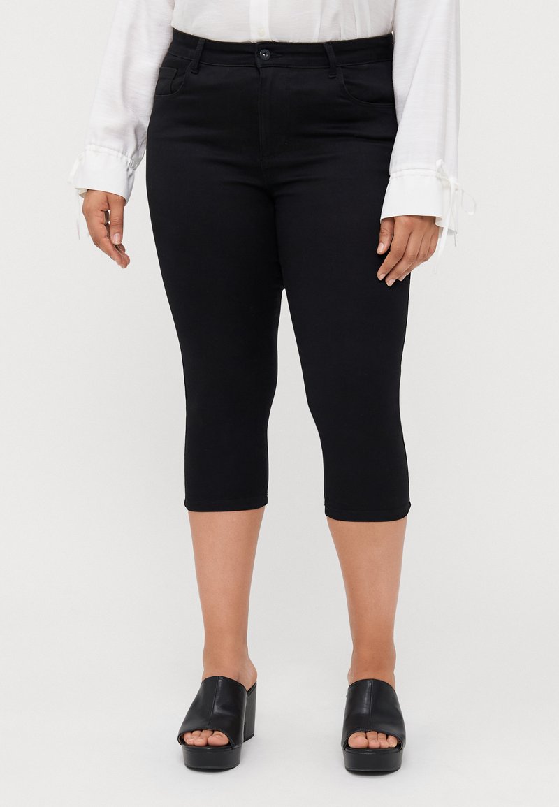 Black capri pants with a slim fit, stretch fabric, and a button closure. Worn with a white long-sleeve shirt and black slide sandals.