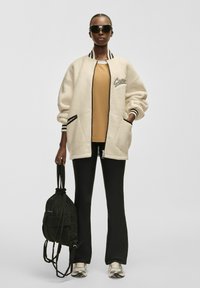 Överdimensionerad krämfärgad fleecejacka med randig svart kant, beige topp, åtsittande svarta byxor och en svart dragsko-väska. Modellen bär solglasögon.