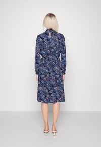 Robe fleurie bleu marine à manches longues, taille cintrée et détail œil-de-perdrix dans le dos. Présente un mélange de motifs floraux colorés.
