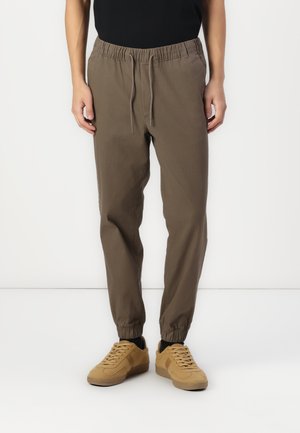 Brown, elastic waist jogger pants with drawstring. Tapered fit, elastic cuffs, and smooth fabric. Paired with tan sneakers.