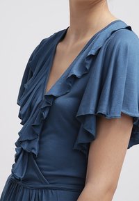 Niebieska sukienka z głębokim dekoltem w kształcie V, dużymi falbaniastymi rękawami i gładką teksturą. Materiał ma miękkie wykończenie z marszczeniami w talii.