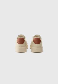 Tan leren sneakers met een beige zool, voorzien van een bruine hexagonale hakaccent en contrasterende stiksels. Minimalistisch ontwerp met ronde veters.