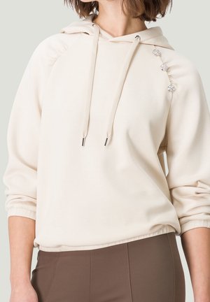 Nainen pukeutuneena kermaiseen huppariin, jossa on kolme koristeellista kristallinappea olkapäällä, ja ruskeat istuvat housut.