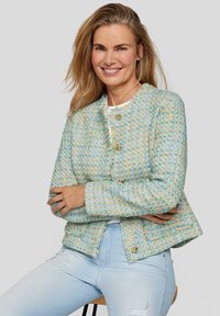 Blaue und gelbe Tweedjacke mit rundem Kragen, goldener Knopfverschluss, strukturiertem Stoff und fransigem Saum. Über einem weißen Oberteil getragen.