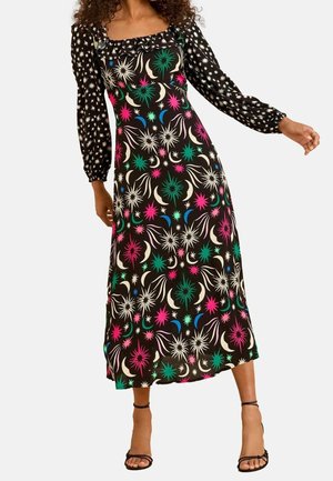 Nainen, joka pukeutuu mustaan midimekkoon, jossa on pitkät tähtikuvioiset hihat ja värikäs taivaallinen kuvio, yhdistettynä mustiin remmikorkokenkiin.