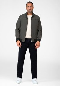 Donkergroene bomberjack met een rits, grijze trui, zwarte jeans en witte sneakers. Glad oppervlak, geribbelde manchetten en staande kraag.