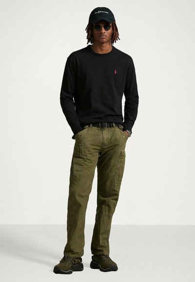Cammicia nera a maniche lunghe, pantaloni cargo verdi con molte tasche, berretto nero e occhiali da sole; calzature casual completano l'outfit.