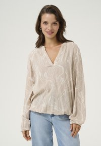 Blouse beige clair avec un col en V, des manches longues et une coupe fluide. Présente un motif abstrait blanc subtil et un tissu doux et texturé.