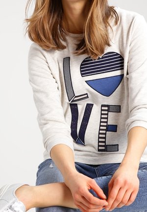 Frau mit hellbraunem Haar, die einen weißen Pullover mit einem blauen, gestreiften Herz und dem Wort "LOVE" trägt, sitzt im Schneidersitz in blauer Jeans und weißen Turnschuhen.