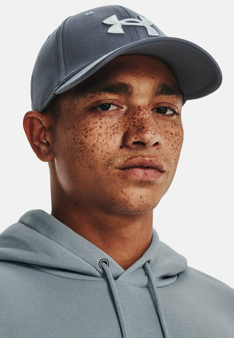 Cappellino grigio con visiera curva, caratterizzato da un logo bianco sul davanti. Chi lo indossa ha una carnagione lentigginosa e indossa un felpa leggera blu.