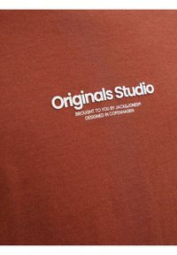 Rūdžių spalvos marškinėliai su lygiu paviršiumi, apvalia iškirpte ir balta spauda, kurioje parašyta "Originals Studio" bei prekės ženklo detalės.