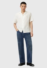 Chemise en lin blanche à manches courtes avec un devant à boutons, assortie à un jean en denim bleu foncé ample et des chaussures noires.
