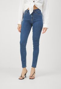 Smal blå denimjeans med hög midja och kontrasterande sömmar. Kombinerat med svarta remsandaler med klack och en knuten vit blus.