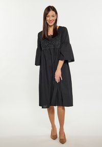 Rochie neagră cu un corset brodat cu flori și mâneci clopot, având o croială lejeră și o lungime până la genunchi, asortată cu pantofi cu toc înalt de culoare bej.
