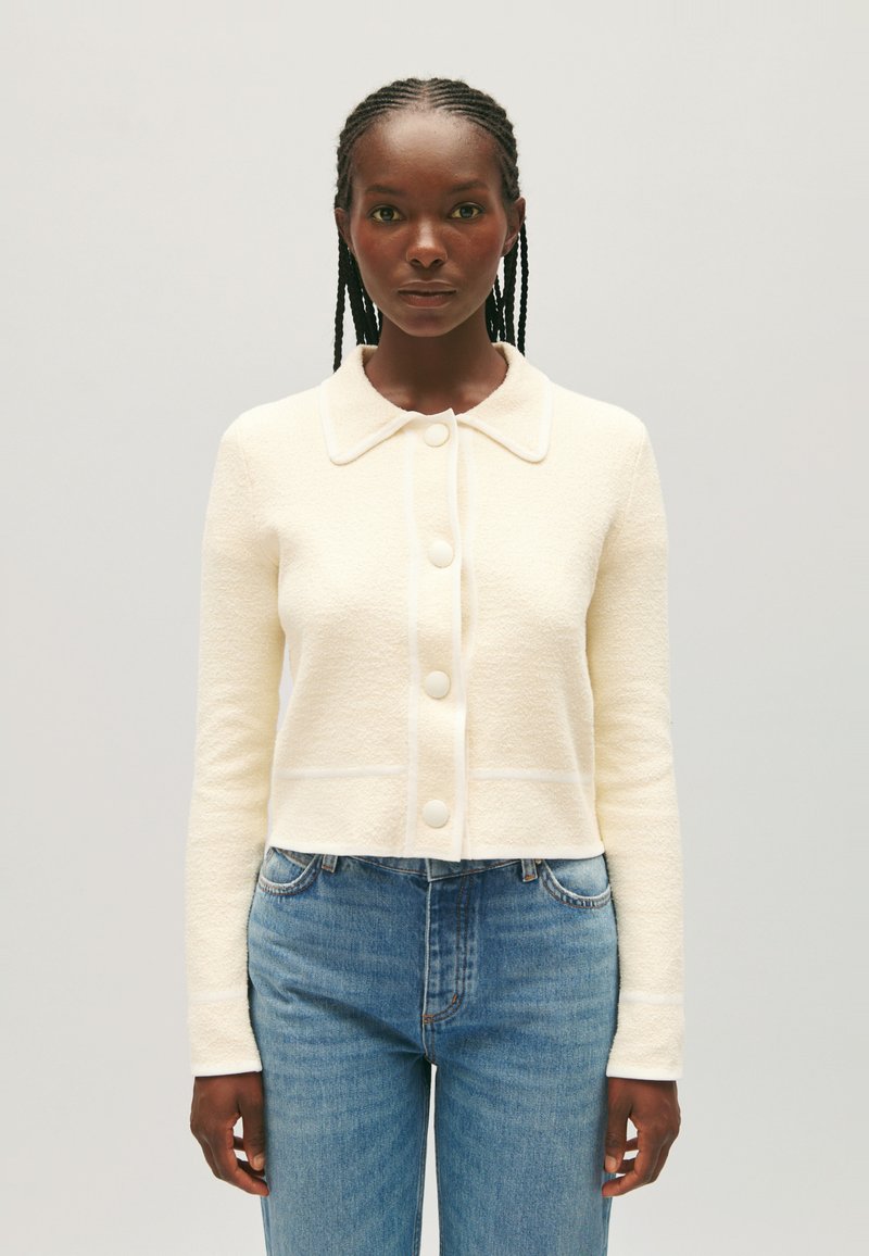 Chaqueta corta color crema con un cuello estructurado, con un tejido texturizado y grandes botones blancos. Combinada con jeans azules.