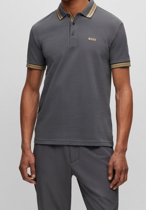 Mężczyzna w ciemnoszarej koszulce polo z krótkim rękawem, z złotym wykończeniem na kołnierzyku i rękawach oraz logo "BOSS" na lewej piersi.