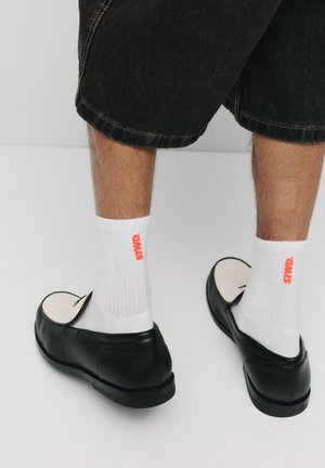 Čierne kožené poltopánky na slip-on s plochou podrážkou, nosené s bielymi rebrovanými ponožkami s červeným nápisom "STWD". Viditeľný lem tmavých denimových šortiek.