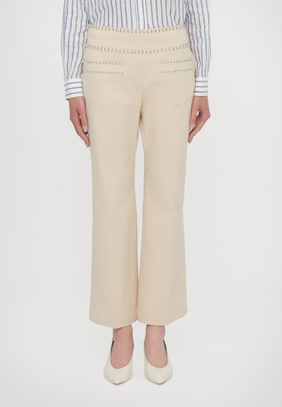 Pantalones de bota acampanada de color beige con acentos de tachuelas en la cinturilla y los bolsillos, hechos de una tela suave, combinados con zapatos de punta crema.