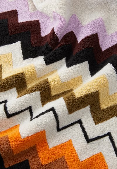 Flerfarget teppe med zigzag-mønster laget av myk, plysj stoff. Har seksjoner i lilla, svart, brunt, gult, oransje og hvitt.