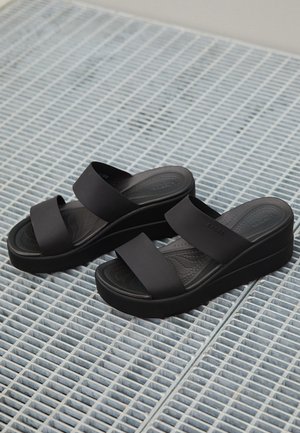 Paire de sandales noires à plateforme avec deux larges brides reposant sur une surface en grille métallique bleu clair.