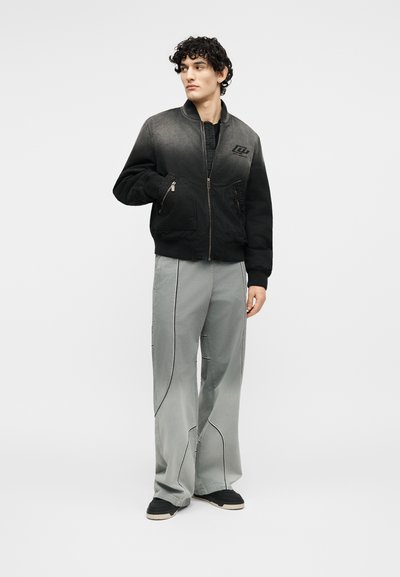Νεαρός άνδρας που φοράει μαύρο μπουφάν μπόμπερ με διαβάθμιση χρώματος και φαρδιά γκρι παντελόνια με μαύρα ραφές, στέκεται με το ένα χέρι στην τσέπη, κοιτάζοντας προς τα δεξιά.