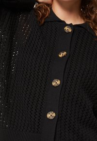 Svart stickad cardigan med ett strukturerat mönster, fem stora, rundade knappar i svart och tan, samt krage med rundad form.