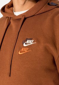 Sudadera Nike marrón con una textura suave. Presenta logotipos multicolores en blanco, naranja y marrón, y una capucha con cordones para ajuste.