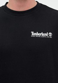Μαύρο μπλουζάκι με στρογγυλή λαιμόκοψη με λευκό λογότυπο Timberland και κείμενο «ΑΝΕΜΟΣ, ΝΕΡΟ, ΓΗ ΚΑΙ ΟΥΡΑΝΟΣ» στην αριστερή πλευρά του στήθους.