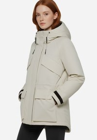 Parka invernale beige chiaro con cappuccio, dotata di molteplici tasche frontali, dettagli neri sui polsini e una texture liscia e opaca.