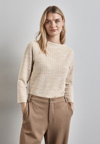 Beige neulepaita, jossa on pyöreä kaula-aukko ja pitkät hihat, yhdistettynä ruskeisiin laskostettuihin housuihin, joissa on nappikiinnitys ja sivutaskut.