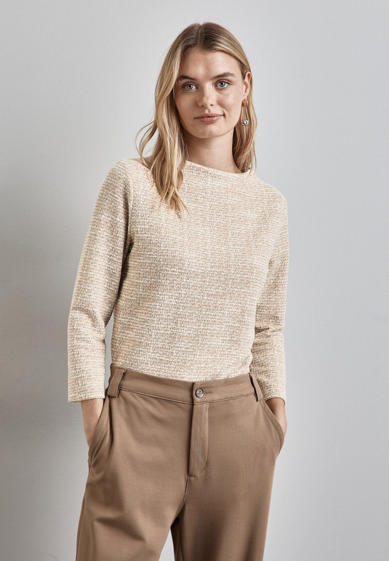 Beige neulepaita, jossa on pyöreä kaula-aukko ja pitkät hihat, yhdistettynä ruskeisiin laskostettuihin housuihin, joissa on nappikiinnitys ja sivutaskut.