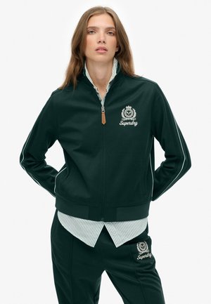 Veste zippée vert foncé avec des rayures blanches le long des bras et un logo. En dessous, il y a une chemise rayée bleu clair et blanche.