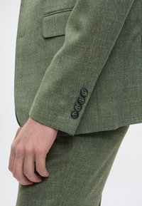 Grüner texturierter Anzug mit einer taillierten Jacke, die eine einzelne Tasche aufweist, ergänzt durch vier schwarze Knöpfe am Ärmel und einer passenden Hose.