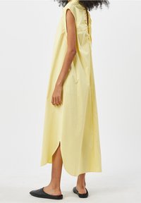 Donna in un abito midi giallo senza maniche e morbido, con uno spacco laterale, in piedi di lato, che indossa scarpe flat nere senza lacci su uno sfondo bianco.