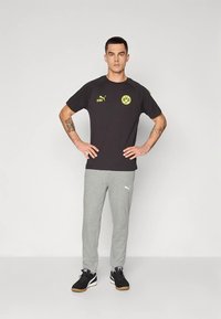 Camiseta negra de Puma con logo amarillo y texto "KING", combinada con pantalones deportivos grises. Zapatillas negras con suela de goma completan el conjunto.
