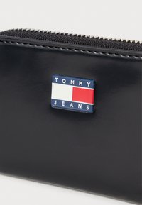 Zbliżenie na czarny skórzany dodatek z srebrnym zamkiem błyskawicznym i prostokątnym logo Tommy Jeans w niebieskim, białym i czerwonym kolorze z przodu.