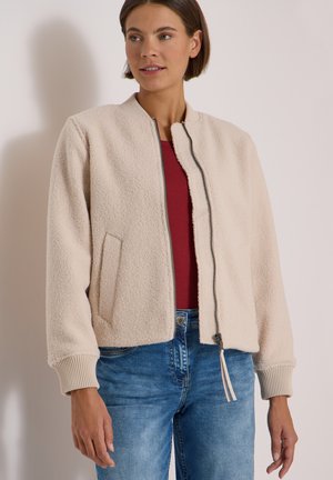 Kvinde iført en beige lynlåset sherpa-jakke over en rød top og blå jeans, stående op ad en ensfarvet lys væg.