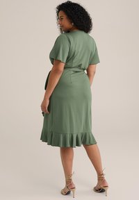 Rochie verde înfofolită cu mâneci scurte, tiv volanat și curea cu legare proprie. Țesătură netedă și design de lungime medie. Sandale bej cu barete completează aspectul.