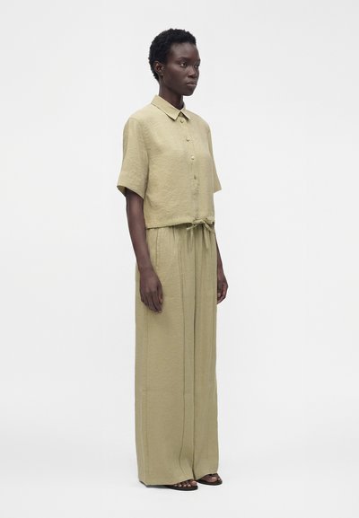 Femme aux cheveux courts et bouclés, portant une chemise beige à manches courtes boutonnée avec ceinture nouée à la taille et un pantalon large assorti, debout de côté.