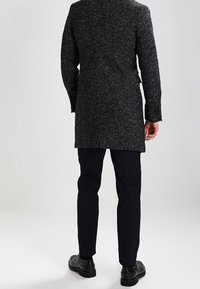 Grå fiskbensmönstrad kappa i texturerat tyg, midjelång modell med slits bak, kombinerad med mörka byxor och skor.