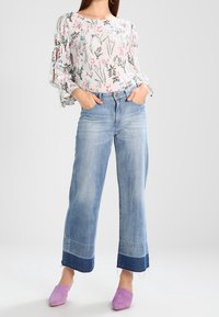 Blusa a fiori con base bianca e maniche arricciate. Jeans a gamba larga di un azzurro chiaro con orlo blu scuro. Scarpe slip-on viola. Sfondo neutro.